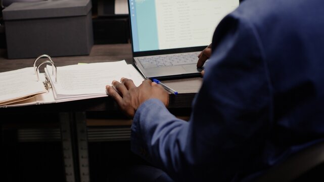 Detective captured in midst of investigation, analyzing records and evidence. Detailed view of policeman at desk with laptop and case files opened for cross-referencing suspect profile and statements.