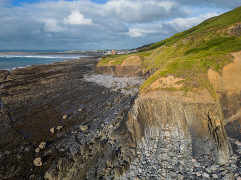 Aerial view towards Westward Ho! town and haunted house. Above dramatic rocky cliff and the beach at low tide with green hill. Sunny day with clouds. Waves of the ocean.