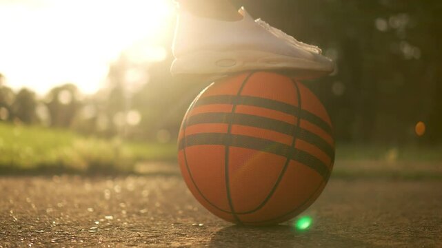 White women sneaker on basketball ball at sunset. Unrecognizable woman places her foot on orange ball in close-up shot with glow of setting sun in background. Sports healthy living and well being