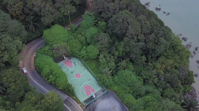 Aerial drone footage of an exotic basketball court by the seaside with a countryside road surrounded by forest. Green nature living. Wellness and wellbeing environment.