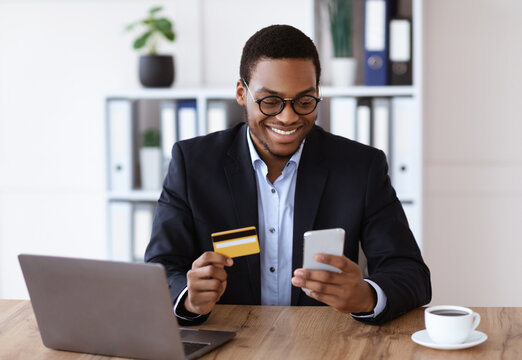 Online shopping, e-payment service concept. Happy african american businessman holding credit card and mobile phone, making e-payments while working with laptop in his office, free space