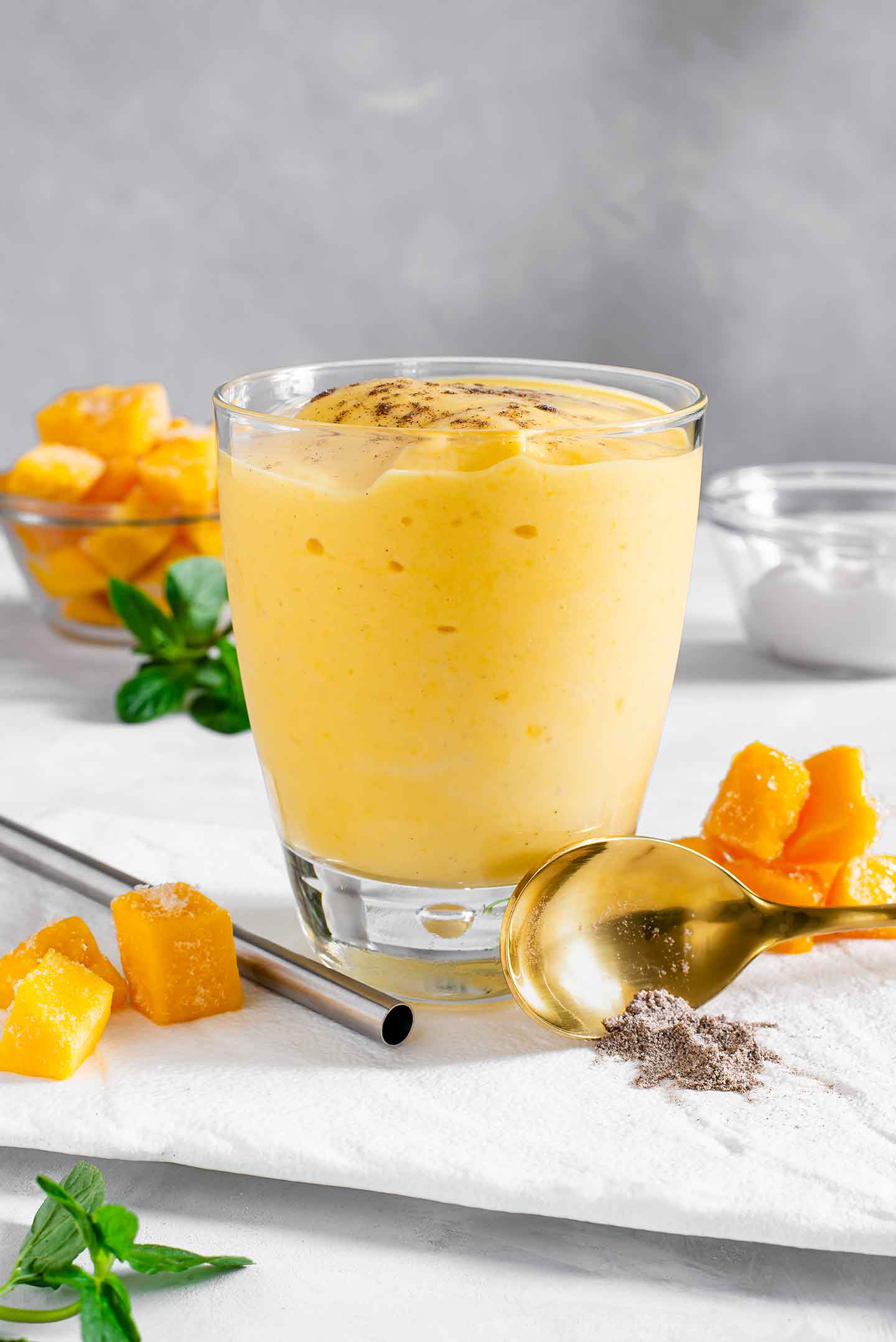 Side view of a thick and creamy mango lassi in a glass with mango cubes, mint leaves, ground cardamom, and a metal straw surrounding the lassi