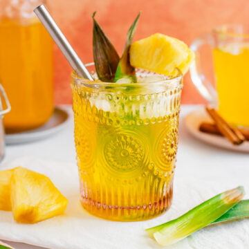 Side view of a cold glass filled with yellow pineapple skin tea. The glass is garnished with a wedge of fresh pineapple and pineapple leaves.