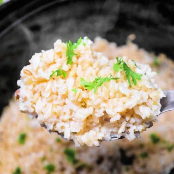 Close-up of a spoonful of slow cooker brown rice garnished with parsley, held over a pot with more rice inside.