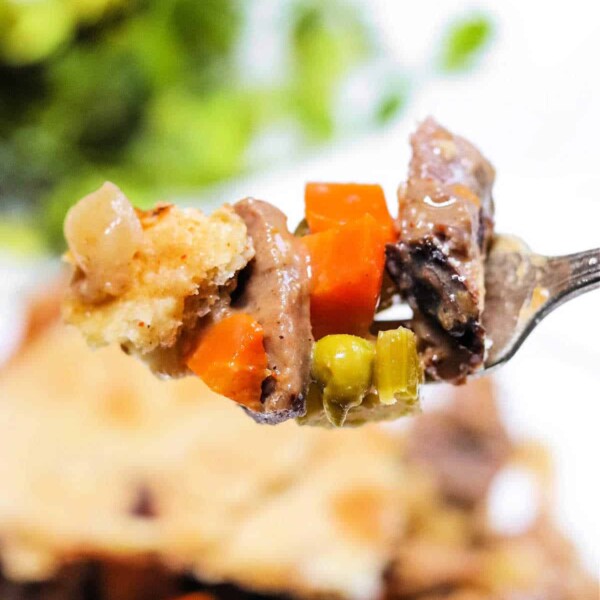 A fork holds a bite of double crust pot pie, showcasing peas, carrots, and crisp golden crust; blurred pie and vibrant greens create a cozy background.