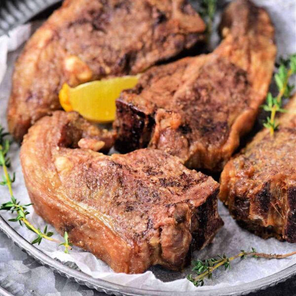 Four perfectly seasoned lamb chops, cooked to tender perfection in an air fryer, rest on a plate adorned with a lemon wedge and fresh thyme sprigs.