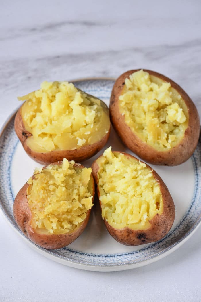 A plate with four halved baked red potatoes. Each potato half has its insides scooped out and slightly mashed but remains in the potato skin. The plate is placed on a white surface.