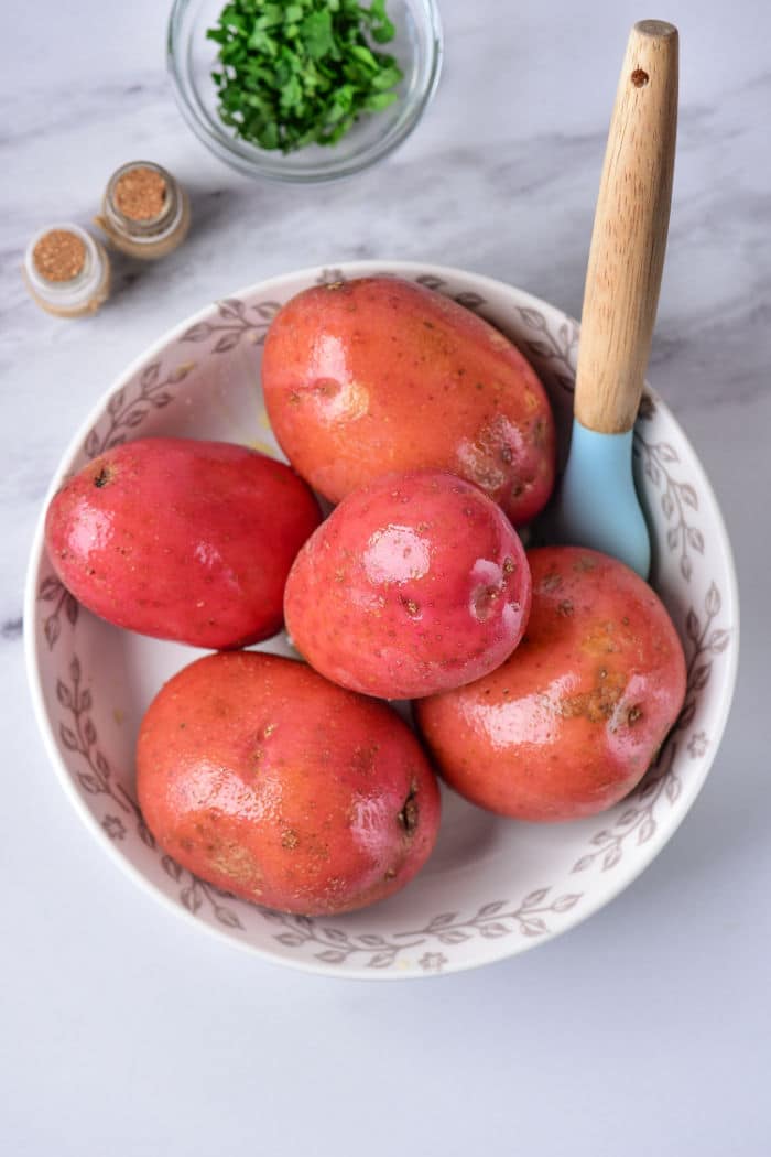 A white bowl with a floral pattern contains five red potatoes and a light blue vegetable peeler with a wooden handle, perfect for prepping baked red potatoes. In the background, on a marble surface, there's a small bowl of chopped herbs and two spice jars.