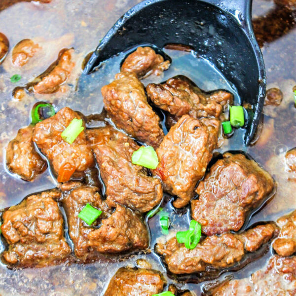 Close-up of braised beef tips stew with succulent chunks of beef, garnished with green onions, in a rich dark brown sauce gracefully cradled by a black ladle.