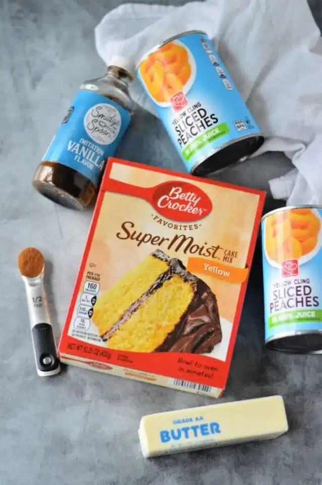 A flat lay of baking ingredients on a gray surface features box cake mix hacks like a Betty Crocker Super Moist Yellow Cake Mix, two cans of sliced peaches, vanilla flavoring, a stick of butter, and a 1/2 teaspoon measuring spoon with spice.