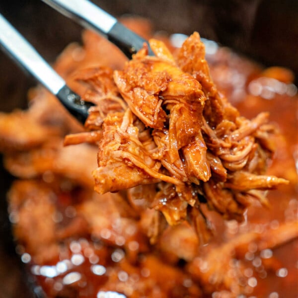 Tongs holding shredded BBQ pulled pork over a slow cooker full of this mouthwatering 2 ingredient pulled pork recipe.