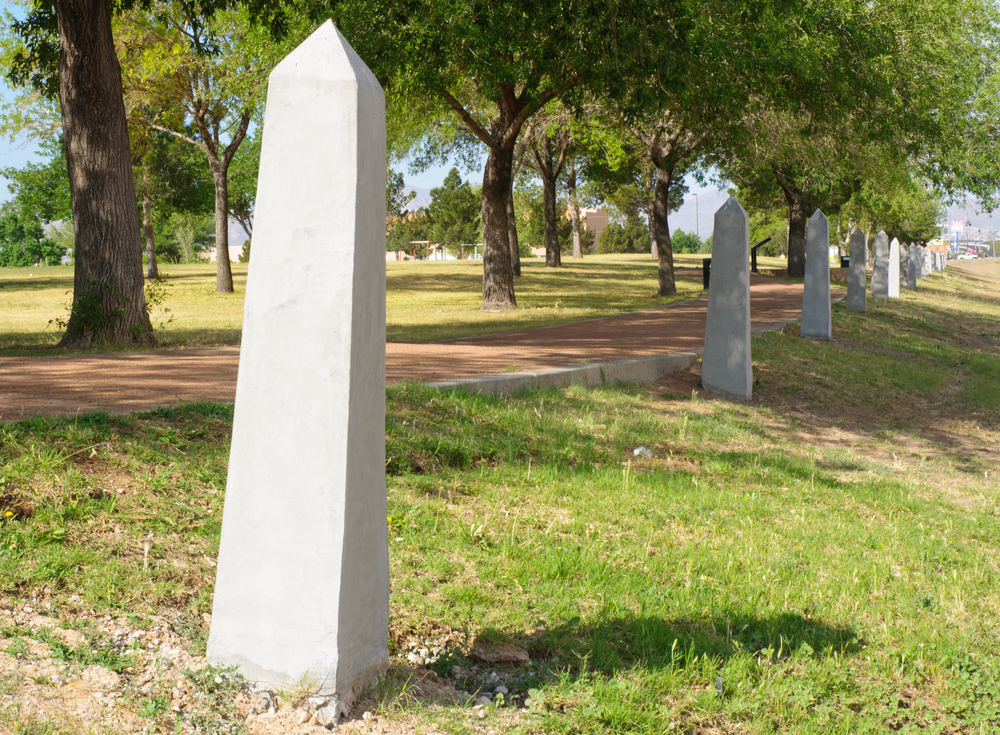 a row of tall pyramid shaped columns in grass shrouded in trees on a sunny day