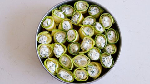 A round baking dish filled with rolled zucchini slices, each containing a creamy white filling with herbs. The zucchini rolls are arranged upright, closely packed together in the dish.