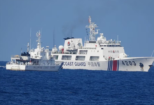 A white Philippine Coast Guard ship is stationed in the waters of the West Philippine Sea, facing a larger Chinese Coast Guard ship. The sea is a deep blue color, and the sky is a dull blue.