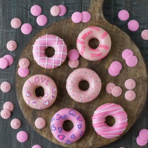 Six Valentine’s Day Donuts on a wooden board surrounded by pink candy melts.