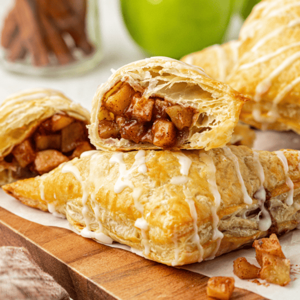 two halves of an apple turnover, showing the inside with apple filling, with half leaning on top of a whole turnover and more turnovers in the background