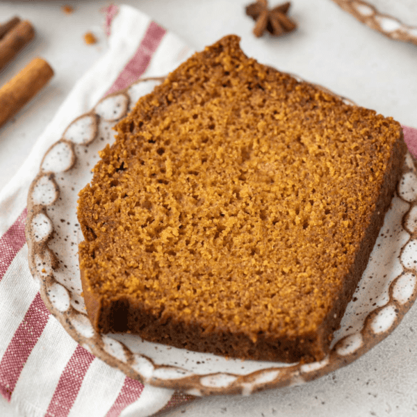 a slice of pumpkin bread on a small plate on top of a red and white towel with cinnamon sticks and star anise scattered around