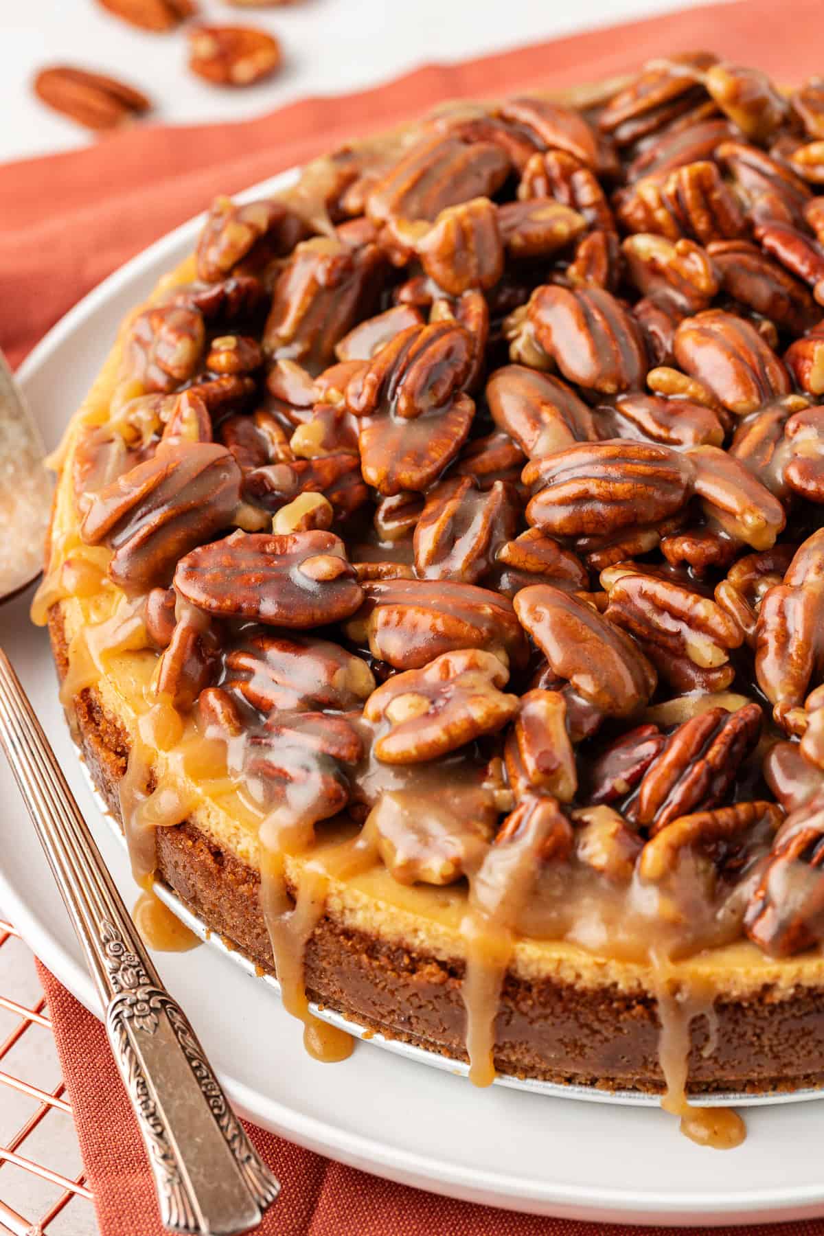 close up of pecan pie cheesecake on a white serving plate on top of a burnt orange cloth on a wire rack with pecans scattered around