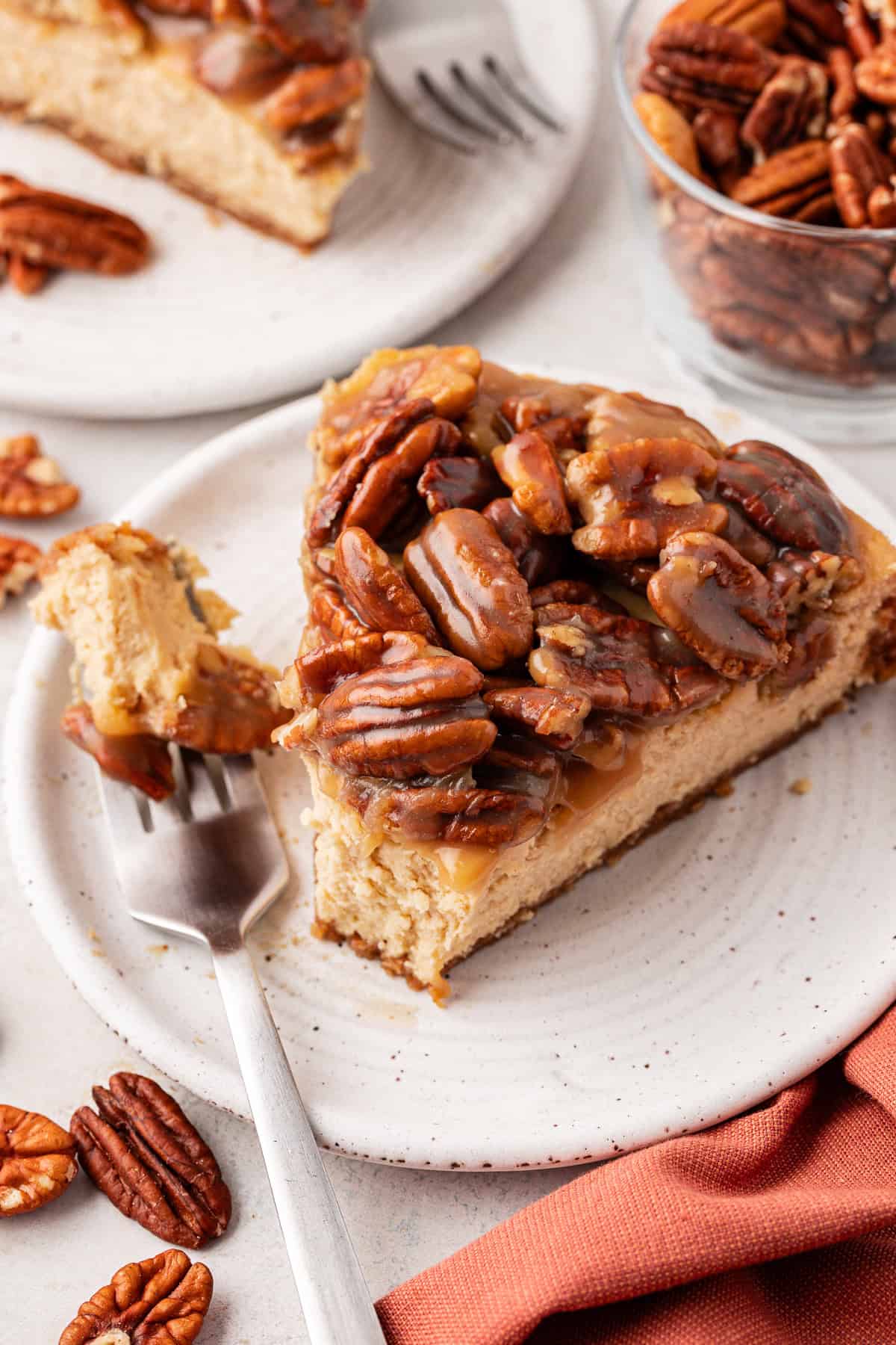 a slice of pecan pie cheesecake on a small white plate with a fork laying beside it with one bite of cheesecake on it, surrounded by a burnt orange cloth, scattered pecans, a glass dish of pecans and another slice on a separate plate