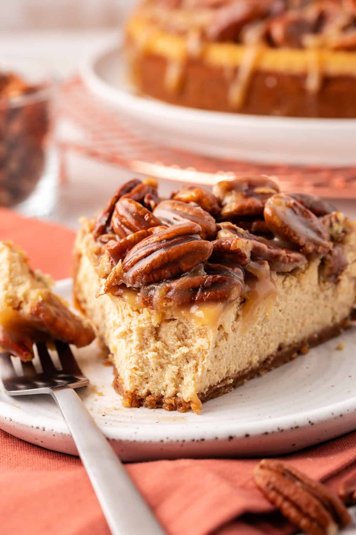 a slice of pecan pie cheesecake on a small white plate with a fork with one bite on it on the side of the plate, on top of a burnt orange cloth with scattered pecans and the whole pie in the background