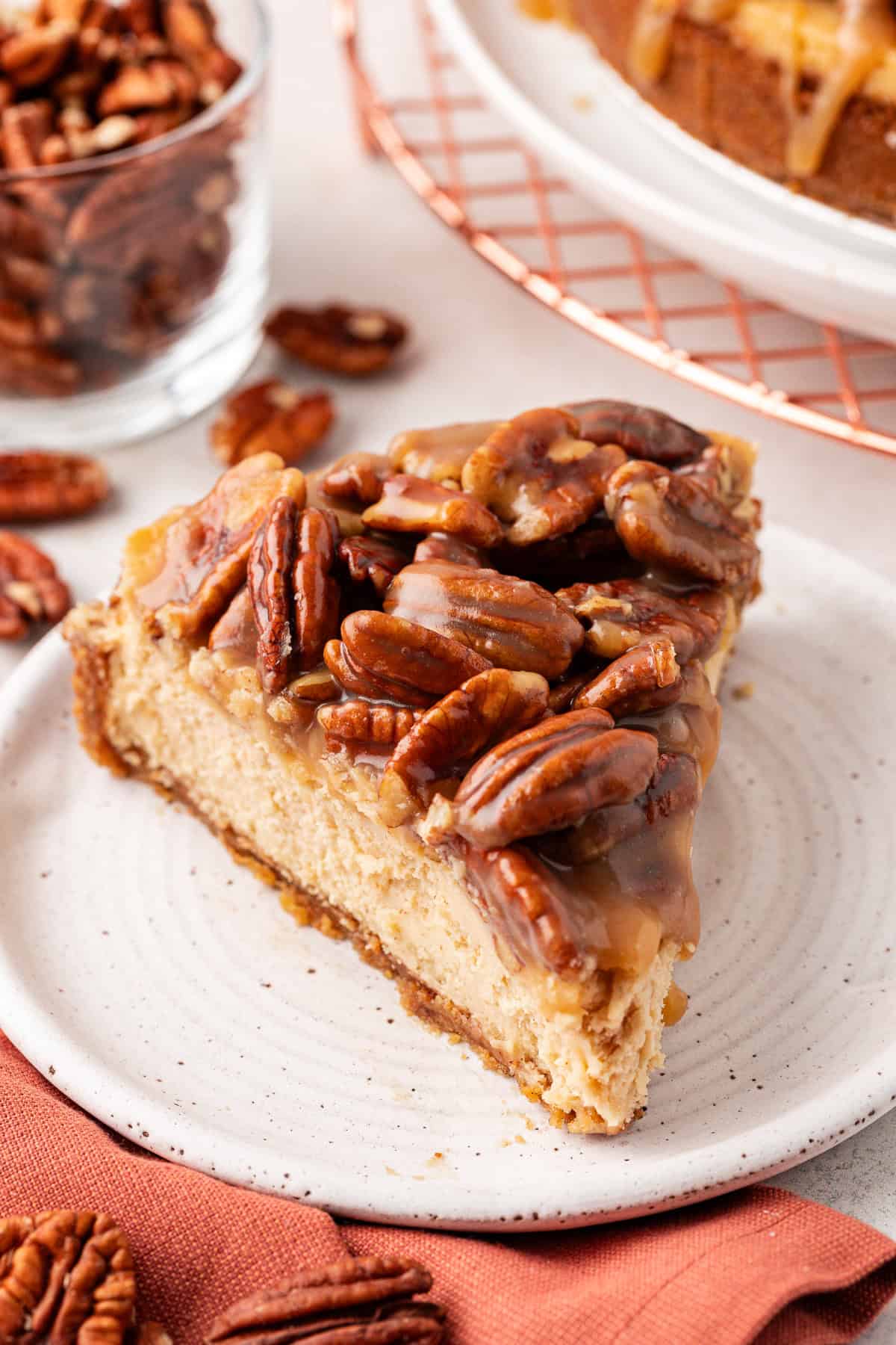 a slice of pecan pie cheesecake on a small white plate beside a burnt orange cloth with scattered pecans around it