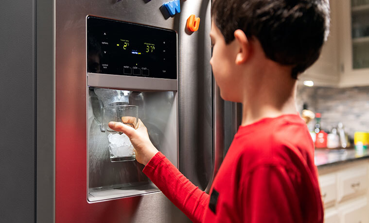 A young boy holds his cup to the iced water dispenser on the door of his fridge.