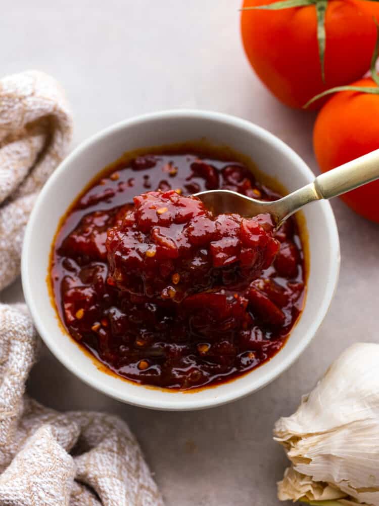 Top view of tomato chutney in a white bowl with a gold spoon lifting up chutney.  A tan kitchen towel and whole tomatoes are next to the bowl of chutney.