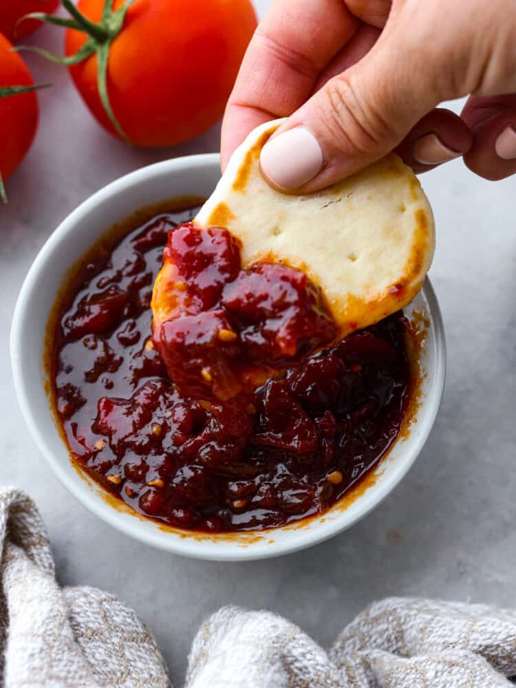 Photo of a piece of naan bread being dipped into a bowl of tomato chutney.  Tomatoes and a kitchen towel are styled next to the bowl of chutney.