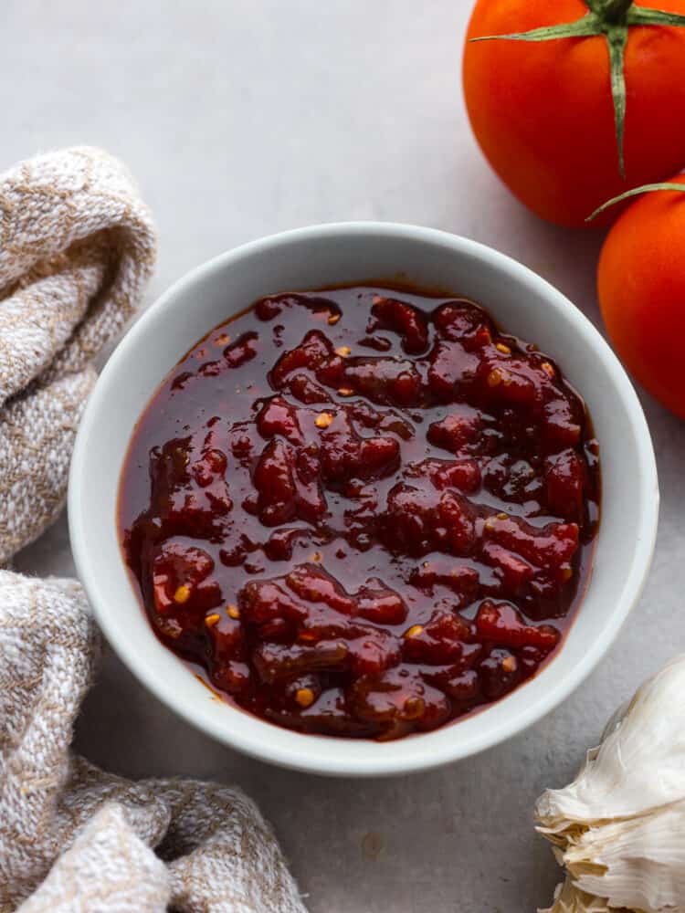 Top view of tomato chutney in a small white bowl with tomatoes and kitchen towel next to the bowl.