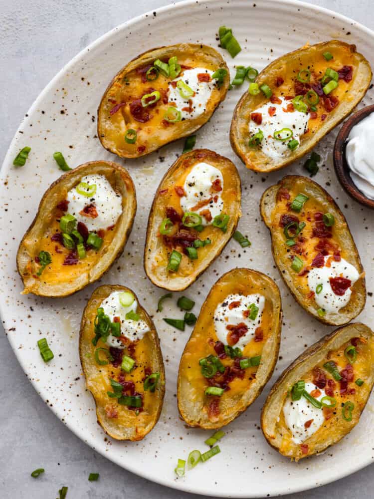 Top view of air fryer potato skins on a large white platter garnished with sour cream, green onions, and bacon. A small bowl of sour cream on the side.