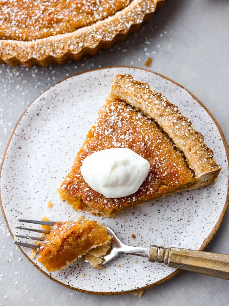 A piece of tart with a scoop sitting on a spoon, topped with whipped cream and the full tart to the side.