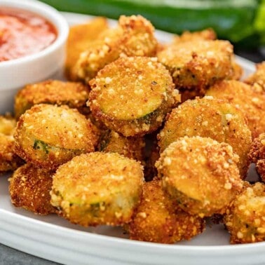 A white platter full of Fried Parmesan Zucchini Chips with a white ramekin full of marinara sauce.