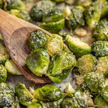 A wooden spoon scoops up Garlic Butter Roasted Brussel Sprouts from a sheet pan.