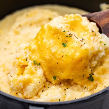 A wooden spoon dipped into a pot filled with cheesy grits.