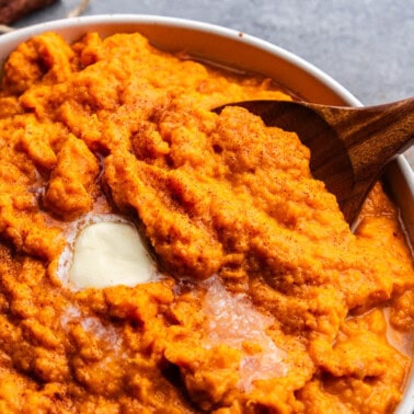 A close up overhead view of a large bowl of cinnamon honey butter mashed sweet potatoes. There's a pat of butter on top that's melting and a wooden spoon set in the bowl ready to serve.