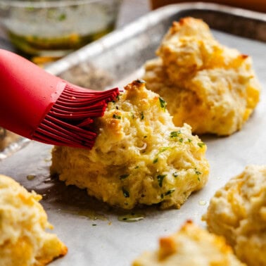 A silicone brush brushing butter onto a cheddar bay biscuit on a pan.