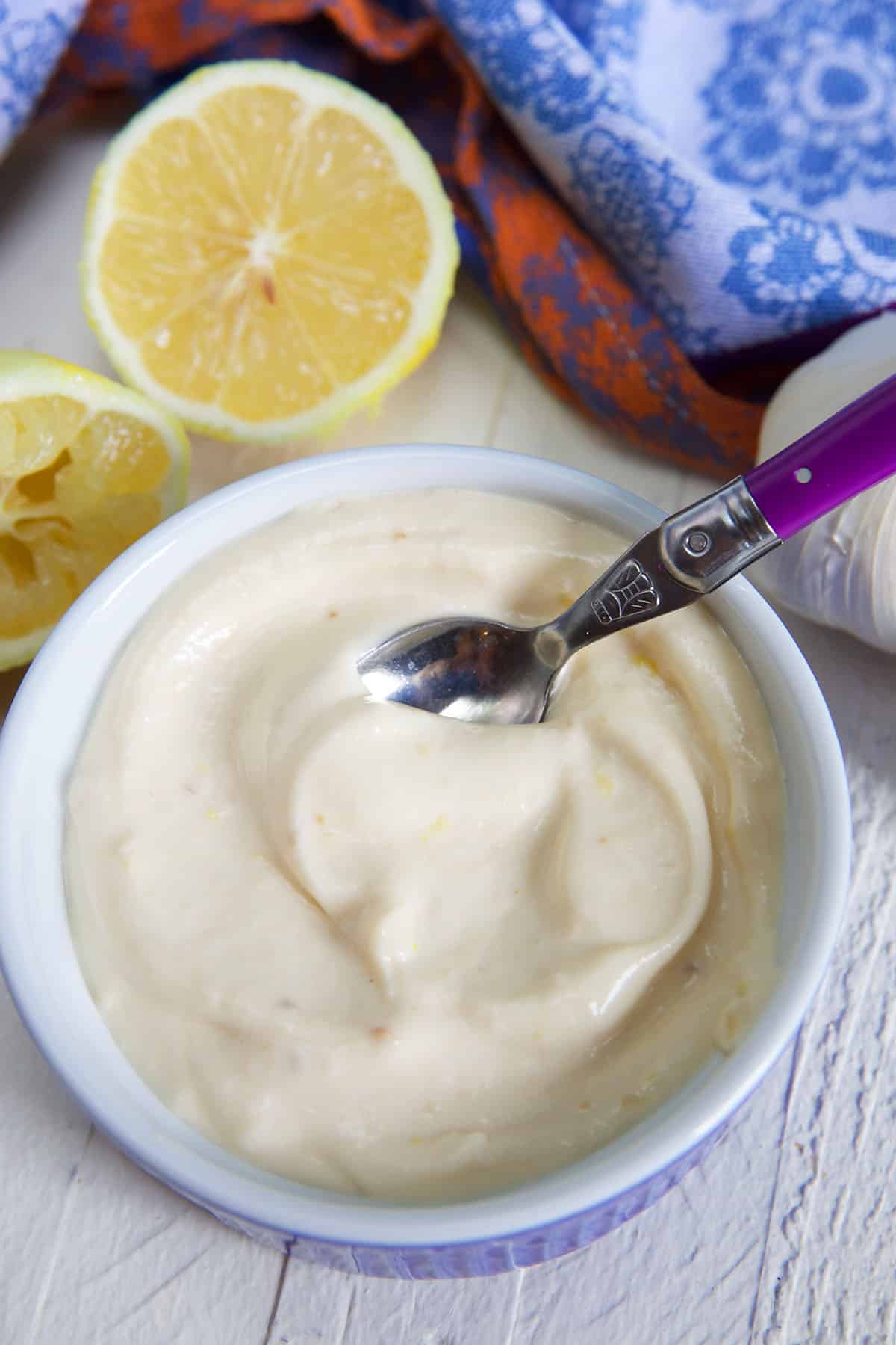 garlic aioli in a blue and white ramekin with a purple handled spoon in it and a sliced lemon on the side.