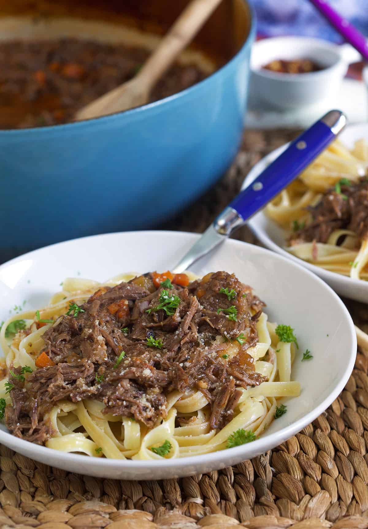 A blue fork is placed in a white bowl filled with noodles and short rib ragu.