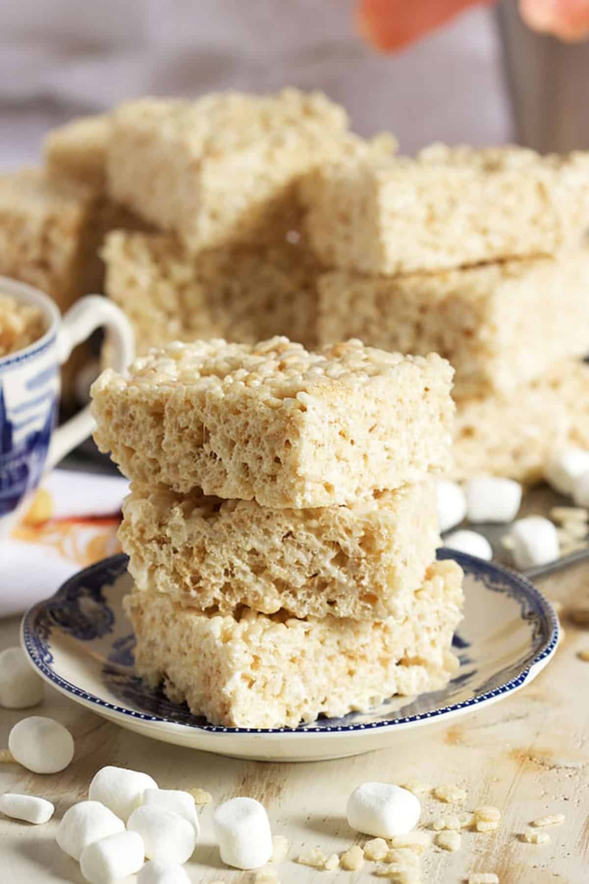 Three Rice Crispy Treats stacked on a blue and white saucer with mini marshmallows in front.