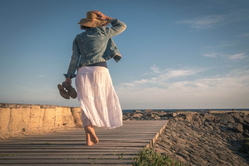 Woman in a denim jacket and broomstick skirt walking barefoot on a wood plank deck. 