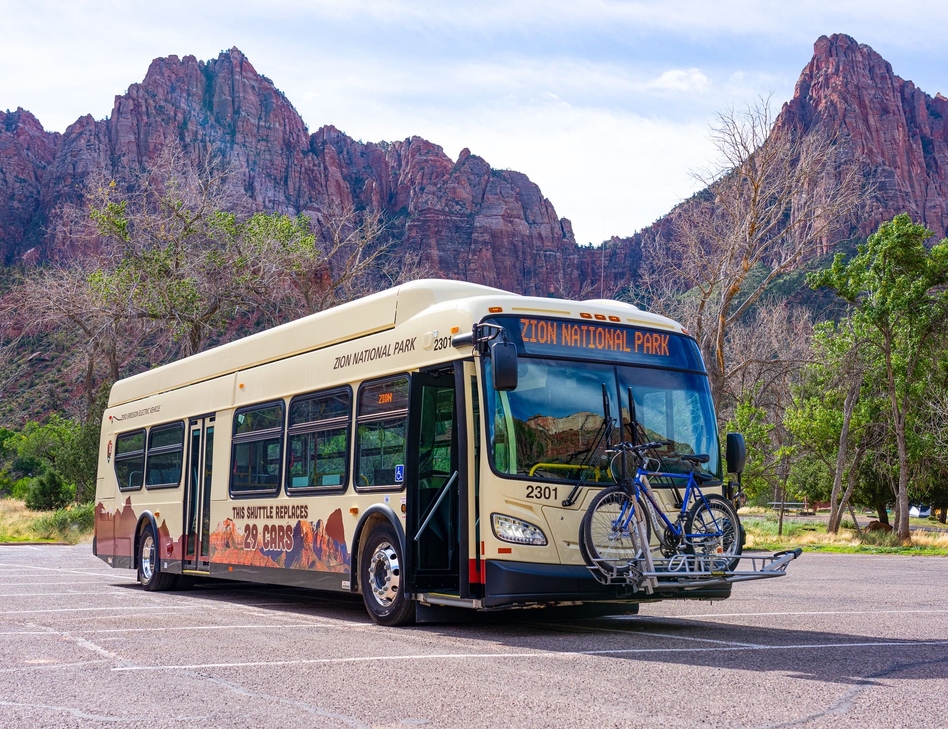 Electric shuttle bus with bikes on a rack mounted in front stands in Zion National Park with red rock towering behind. Text on bus reads, "This shuttle replaces 29 cars." A new electric shuttle bus in front of The Watchman in Zion National Park.