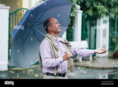 Happy middle-aged man with umbrella outstretching hand to catch rain ...