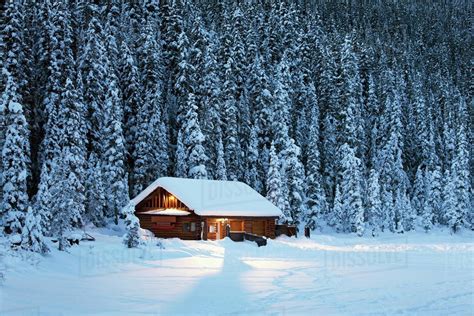 A snow covered log cabin on a snow covered lakeshore surrounded by ...