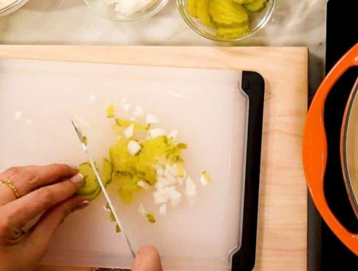 Manicured hands chopping pickles and onions on a white cutting board.