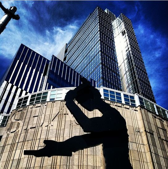 Silhouette of a large sculpture of a man tipping his hat in front of a modern high-rise building against a bright blue sky. The sculpture is positioned in the foreground, giving a dramatic contrast to the glass architecture behind it.