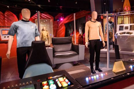 Mannequins dressed in iconic blue and beige uniforms stand in a sci-fi themed exhibit, featuring a chair and various control panels with colorful buttons, evoking a classic starship bridge setting.