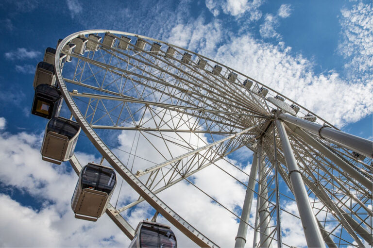 A low-angle view of a large Ferris wheel with several gondolas against a blue sky filled with fluffy white clouds captures one of the must-see things to do in Seattle. The perspective emphasizes the wheel's structure and the impressive height of this iconic amusement ride.