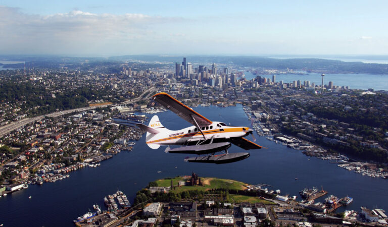 A seaplane glides over Seattle, with a stunning view of downtown skyscrapers in the distance. Below, the cityscape offers a mix of buildings, roads, and the shimmering Puget Sound surrounded by lush green shorelines. The sky is clear—a perfect day to explore endless things to do in Seattle.