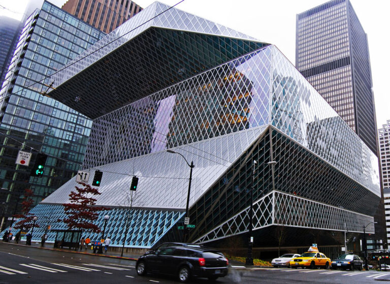 A modern, angular building in Seattle boasts a unique geometric glass facade with diamond-shaped patterns. Cars and traffic lights fill the foreground, while towering skyscrapers surround it on a cloudy day, making it an architectural highlight among things to do in Seattle.