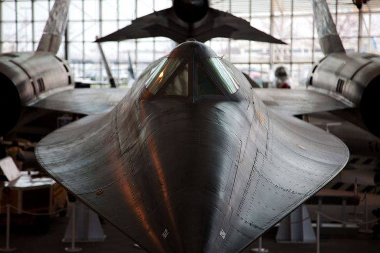 A front view of a sleek, black jet aircraft with sharp angles and a pointed nose is displayed indoors, reminiscent of one of the thrilling attractions on a list of things to do in Seattle. The plane's streamlined design suggests high-speed capabilities against the backdrop of the large window.